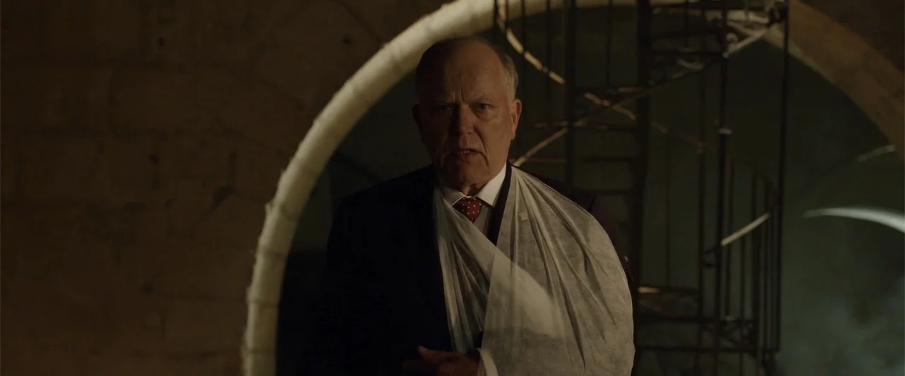 Hounds of War (2024) movie still: medium — A stone cellar features arched walls and a metallic spiral staircase in the background. A man in a suit…