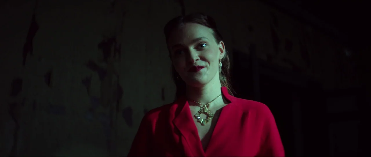 Braid (2018) movie still: medium — A woman stands in a darkened space with indistinct, rough-textured walls behind her. She faces forward wearing a…