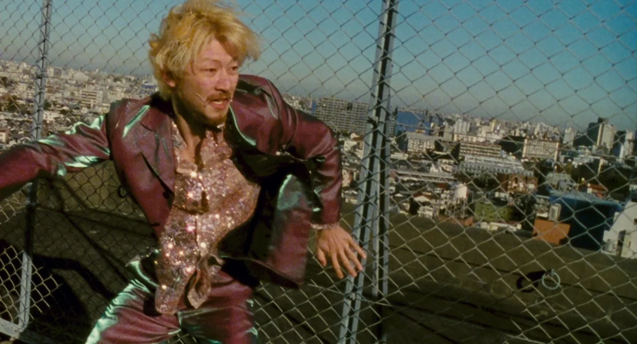 Ichi The Killer (2001) movie still: medium — A man runs along a metal chain-link fence atop a high-rise city building. The background shows a dense…