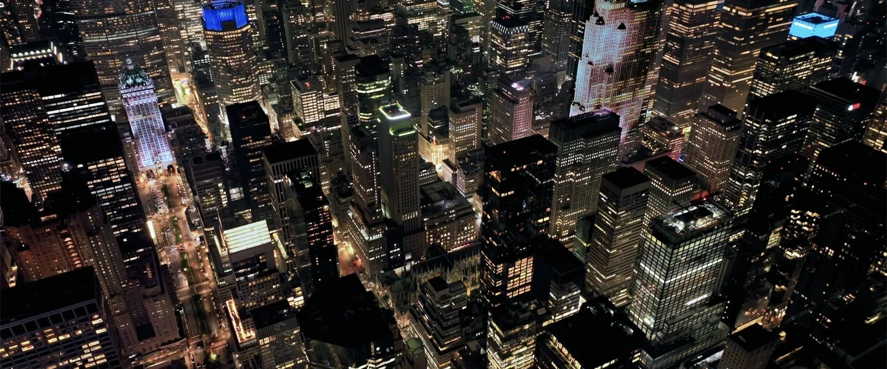 Play Dirty (2025) movie still: establishing — An aerial view of a dense urban grid features illuminated skyscrapers and canyon-like streets. The…