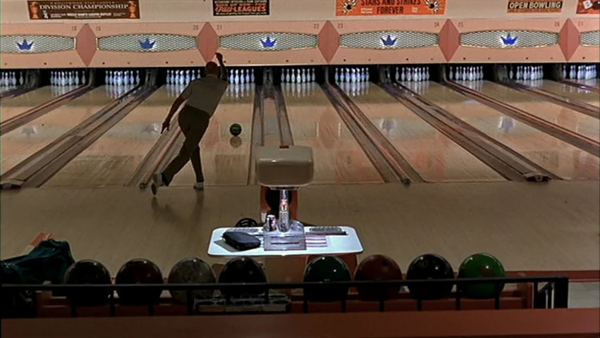 The Big Lebowski (1998) movie still: wide — A classic bowling alley lane features a row of hanging numbered signs above pins. A lone man in a light…
