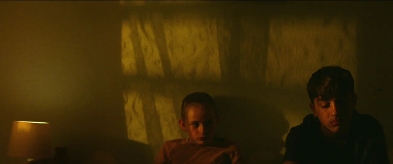 Scrapper (2023) movie still: medium — A dimly lit room with textured walls displays a long shadow cast from a window. A young boy and a girl sit in…
