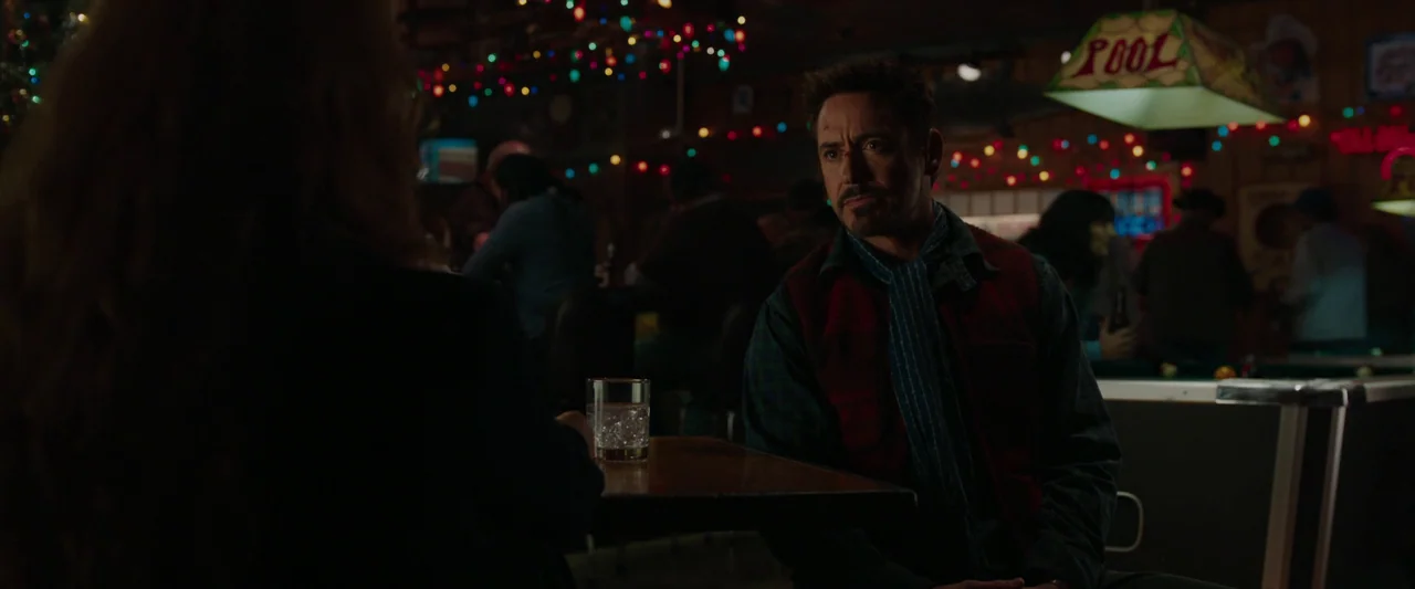 Iron Man 3 (2013) movie still: medium — A dimly lit bar decorated for the holidays with colorful string lights hanging above. A man in a vest sits…