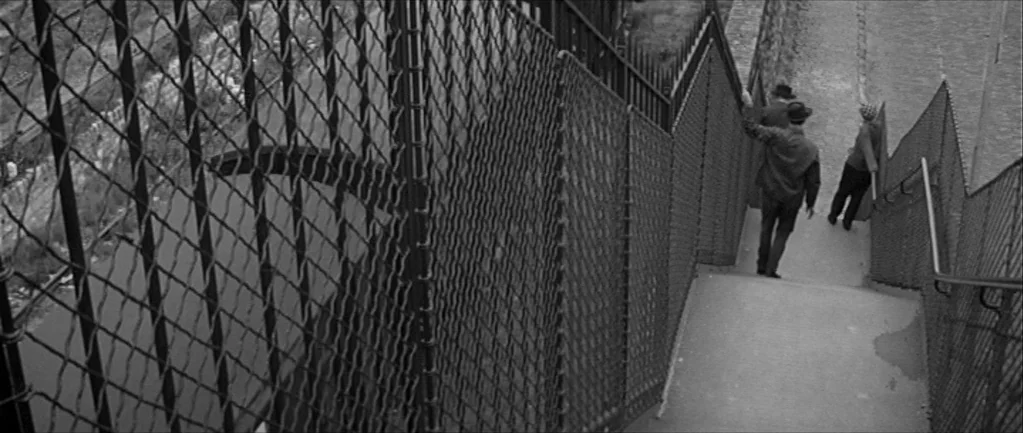 Jules and Jim (1962) movie still: wide — A steep outdoor staircase bordered by tall chain-link security fences and metal railings. Two men and a…