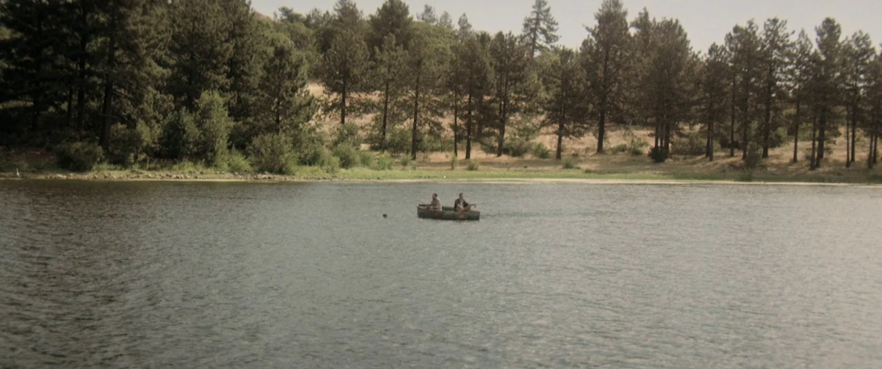 The Endless (2017) movie still: wide — A calm lake is surrounded by a dense line of tall pine trees. Two men sit quietly in a small wooden boat…