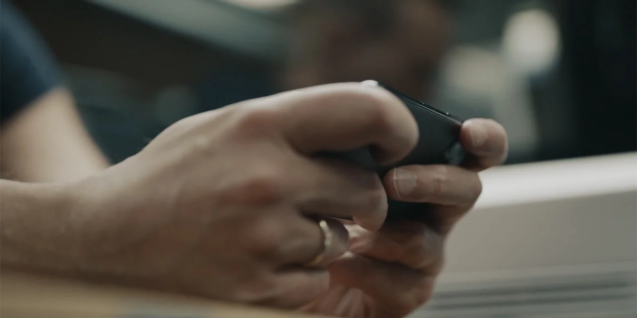 Navalny (2022) movie still: closeup — A close view shows two hands holding a black mobile device inside a transit vehicle. A simple gold wedding…