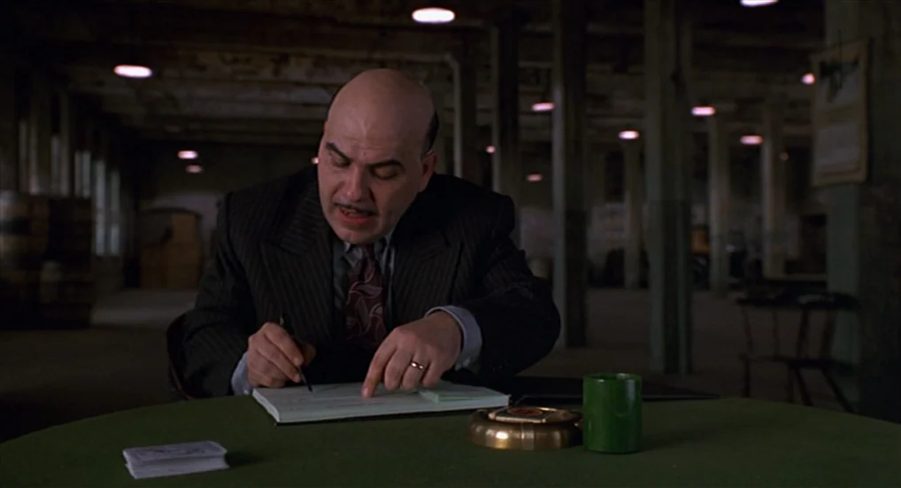 Miller’s Crossing (1990) movie still: medium — A dark, empty warehouse with concrete pillars and overhead industrial lighting. A bald man in a suit…