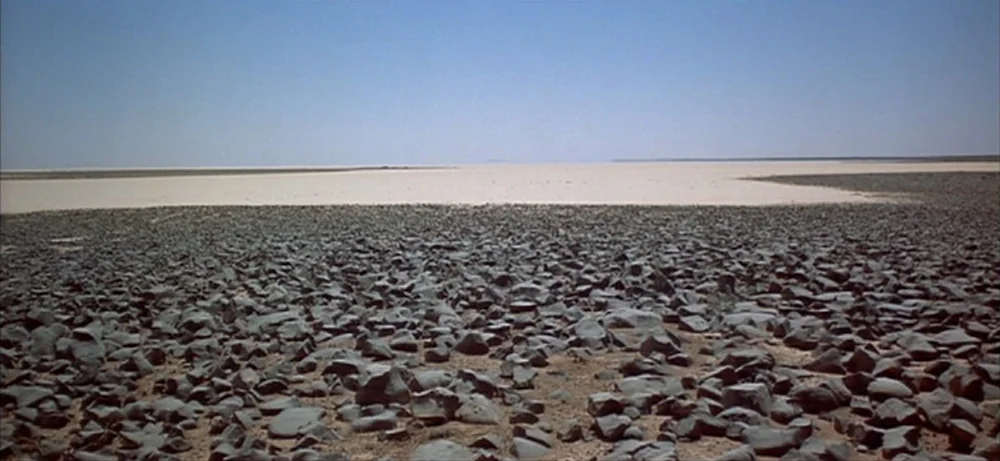 Lawrence Of Arabia movie still: establishing — A vast, flat desert landscape stretches toward a distant, cloudless blue horizon under daylight. The…