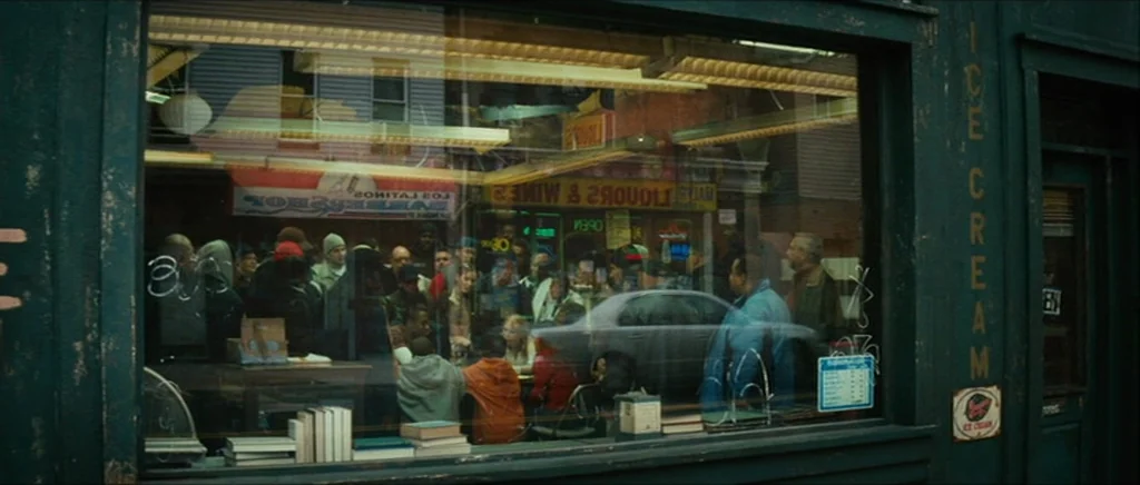 Be Kind Rewind (2008) movie still: wide — A dark green storefront window displays a reflection of a barbershop and liquor store. A crowd of people…
