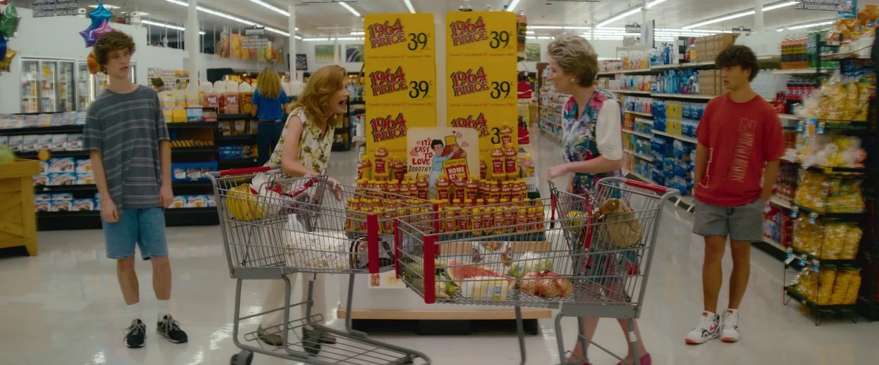 Snack Shack (2024) movie still: wide — A well-lit grocery store aisle features shelves stocked with various colorful consumer products. Two women…