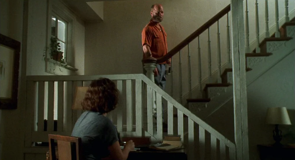 Pollock (2000) movie still: medium — A residential staircase landing with wooden railings and a window overlooking a leafy plant. A man stands on…