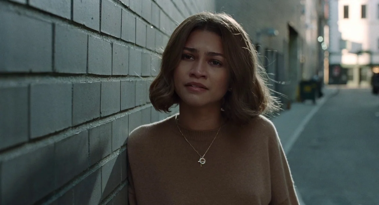 Challengers (2024) movie still: medium — A woman stands beside a gray brick wall in an urban outdoor space. She wears a tan sweater and looks ahead…