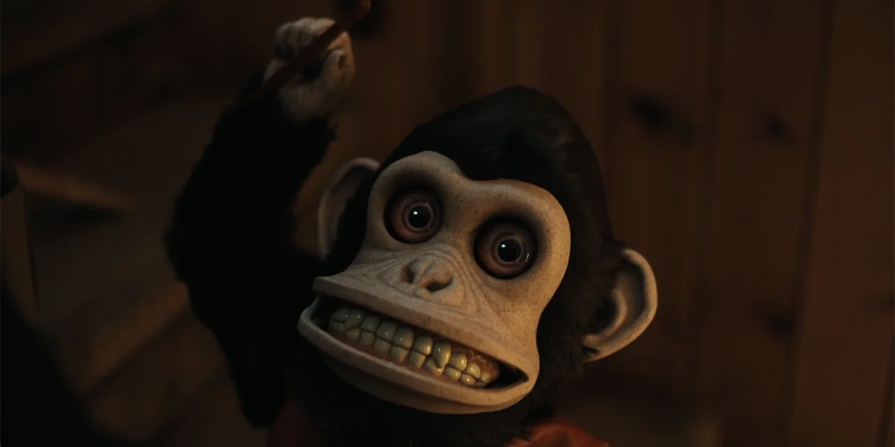 The Monkey (2025) movie still: closeup — A close-up of a textured toy monkey character in a dark indoor space. The monkey is holding a stick in its…