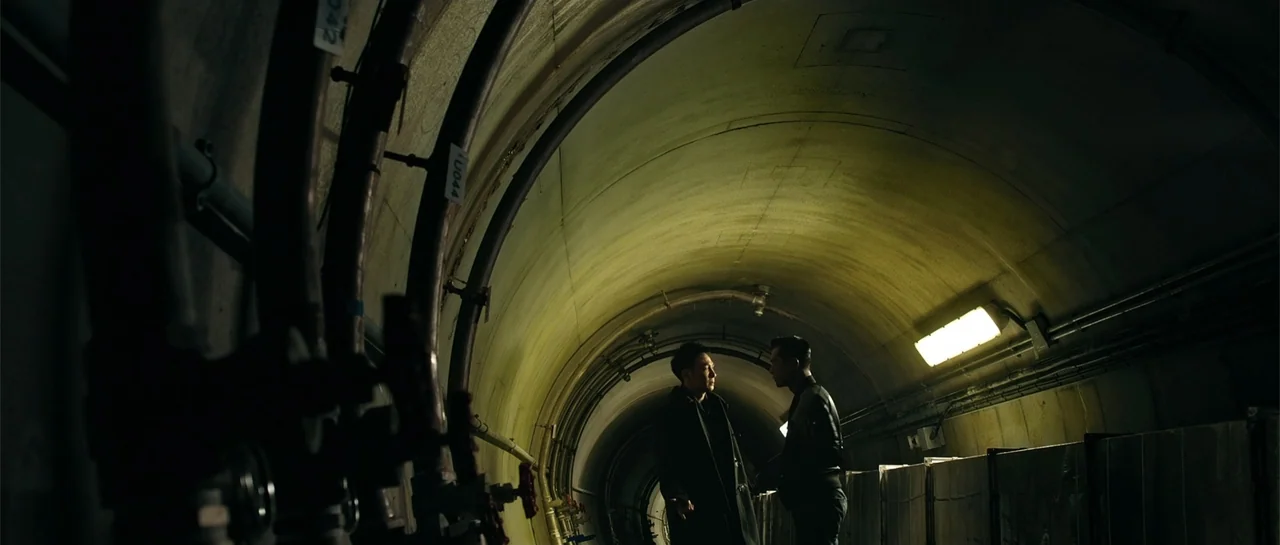 Line Walker (2016) movie still: wide — A concrete utility tunnel with curved walls features exposed piping along the left side. Two men stand facing…