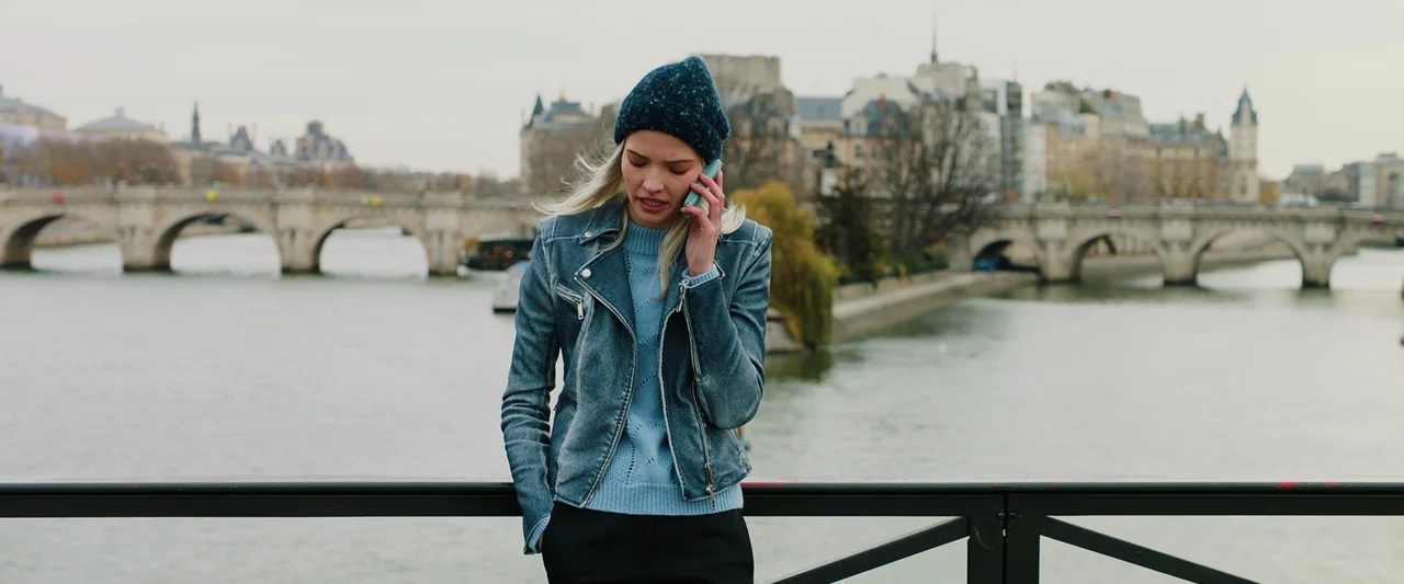 Anna (2019) movie still: medium — A woman stands on a bridge over a river with a city cityscape behind. She looks down while holding a mobile phone…