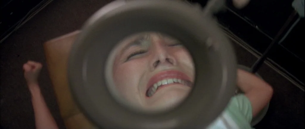 Wild At Heart (1990) movie still: closeup — The frame captures a view through a round dental lamp. A woman lies in a chair with her eyes closed and…
