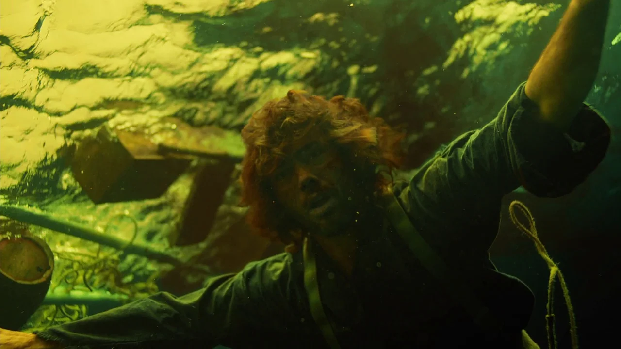 In The Heart of the Sea (2014) movie still: medium — A man drifts submerged in murky yellow-green water near debris and ropes. He reaches his arm…