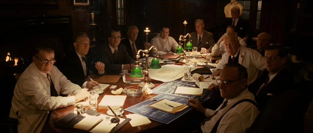 Shutter Island (2010) movie still: wide — A dimly lit mahogany conference room contains a large oval table covered in blueprints. Twelve men in…
