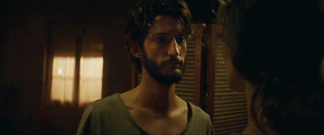 Masquerade (2022) movie still: medium — A dimly lit room features a wooden screen and a window with curtains. A bearded man in a grey t-shirt stands…