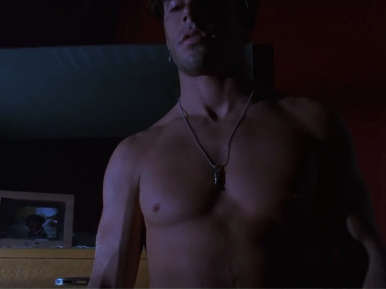 The Living End (1992) movie still: medium — A dimly lit room contains a framed photograph sitting on a wooden surface. A shirtless young man stands…