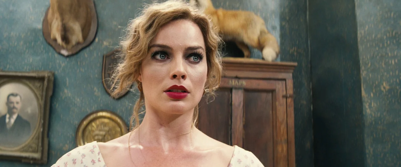 Babylon (2022) movie still: closeup — An interior space with teal walls decorated with taxidermy, framed photos, and wooden storage. A woman stares…