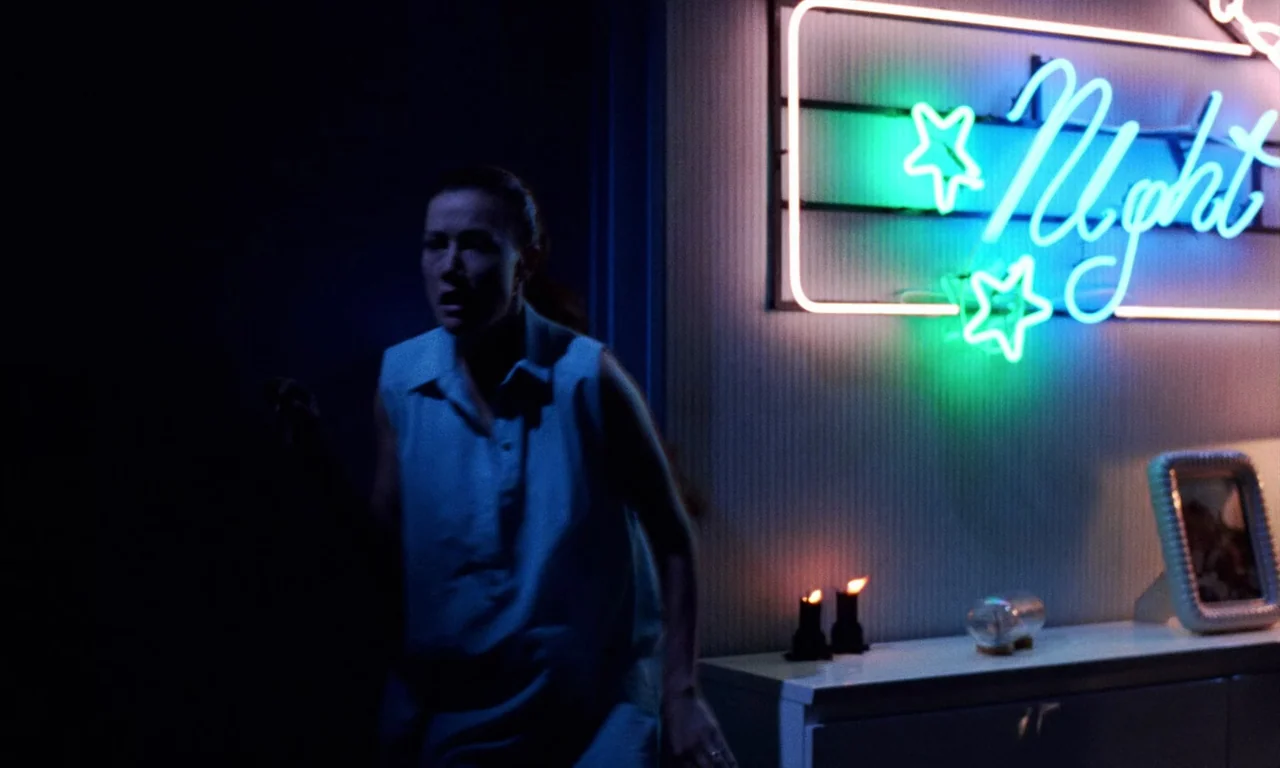 Demons 2 (1986) movie still: medium — A dimly lit room with a neon 'Night' sign mounted on a textured wall. A woman walks through the space past a…