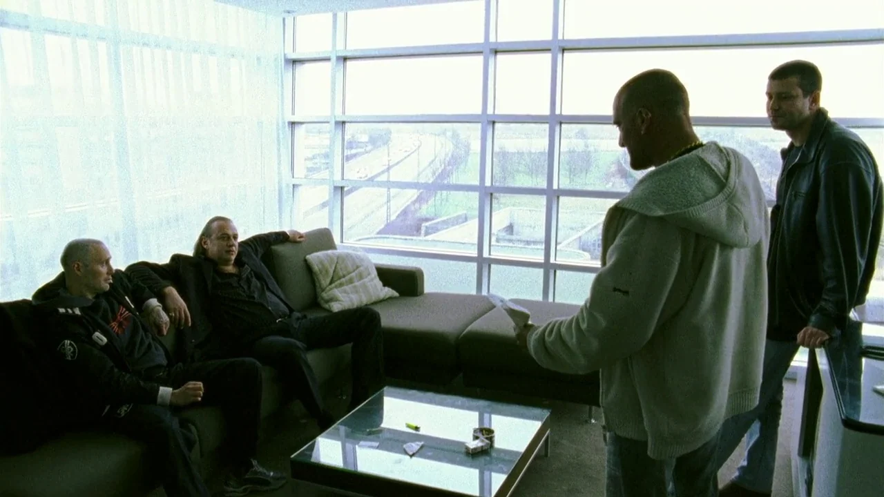 Pusher II (2004) movie still: wide — A modern room features a large window wall overlooking a distant highway landscape. Four men stand and sit on a…