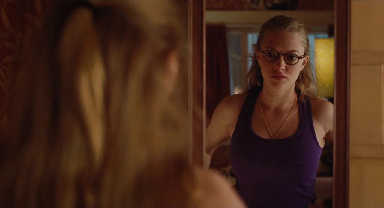 Jennifer’s Body (2009) movie still: medium — A woman stands before a vertical wall mirror in a dimly lit room. She watches her own reflection while…