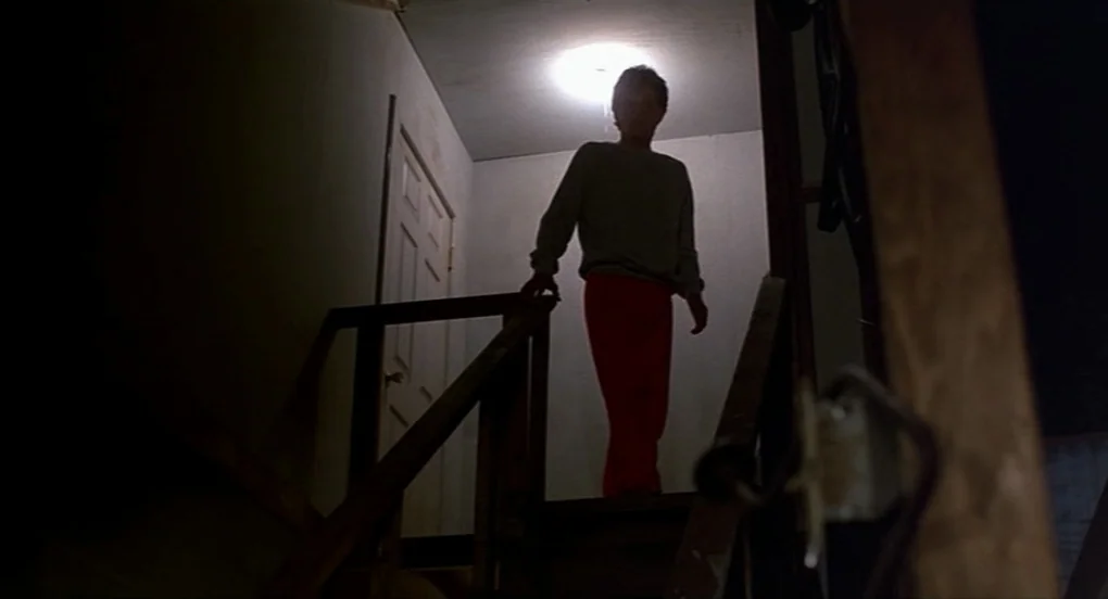 A Nightmare On Elm Street 2: Freddy’s Revenge (1985) movie still: fullbody — A dark staircase leads down from a dimly lit landing with a single bare…
