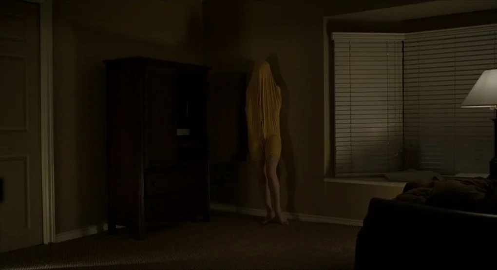 The Future (2011) movie still: wide — A dimly lit room with tan walls features a dark wooden armoire near a window. A person stands in the corner…