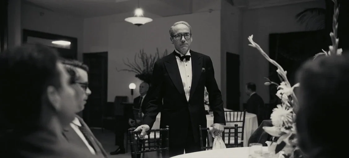 Oppenheimer (2023) movie still: medium — An elegant 1950s formal banquet room with round tables and white tablecloths. A man in a tuxedo stands…