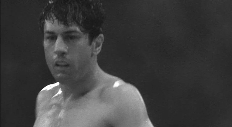 Raging Bull (1980) movie still: closeup — A dark boxing ring illuminated by intense overhead spotlights. A shirtless man looks slightly away with…