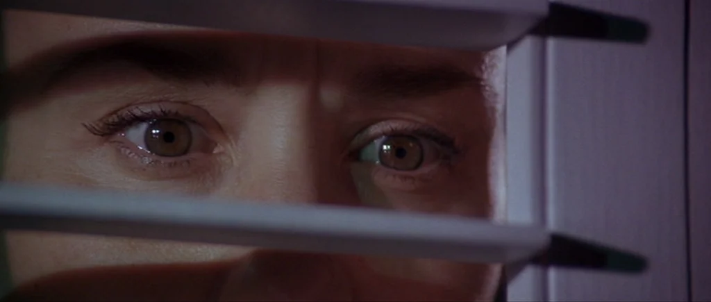 Cape Fear (1991) movie still: closeup — A close view of a person looking through gaps in closed horizontal window blinds. The person stares forward…