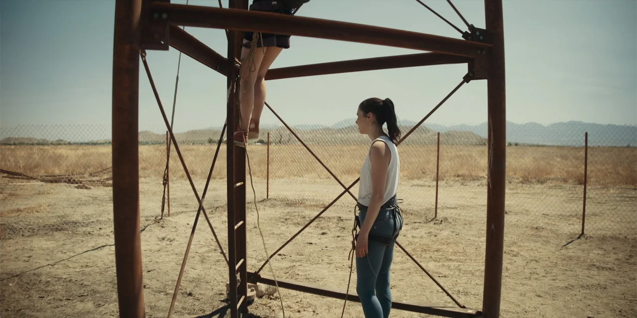 Fall (2022) movie still: medium — A dusty, dry lot features a tall rusty metal structure amidst sparse brush. One woman stands on the ground while…