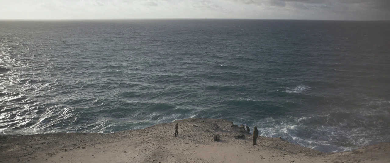 Solo: A Star Wars Story (2018) movie still: wide — A rugged cliff edge overlooks a vast, choppy ocean under a gray clouded sky. Three figures are…