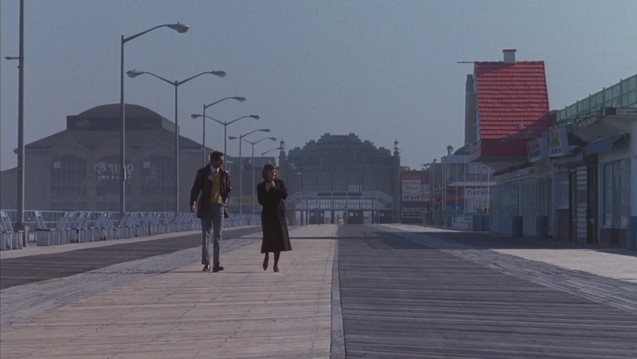 Baby It’s You (1983) movie still: wide — A wide wooden boardwalk stretches into the distance, flanked by buildings and lampposts. A man and a woman…