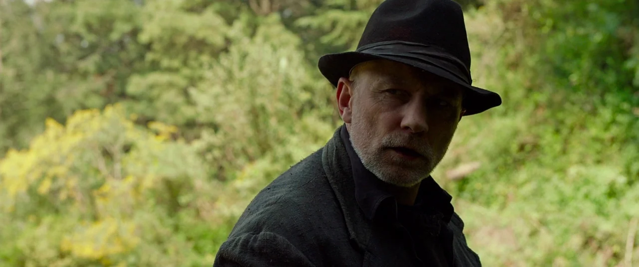 Siberia (2019) movie still: medium — A dense forest with lush green foliage and blurred leafy branches. A man wearing a black hat and dark coat…