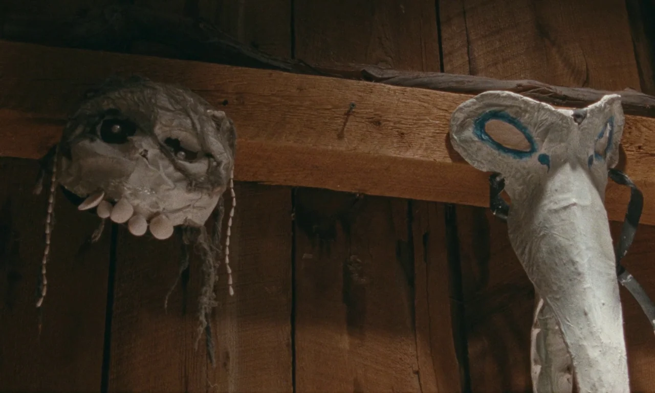 Janet Planet (2023) movie still: closeup — Two handcrafted masks are mounted on a weathered wooden wall inside a rustic building. A grey round mask…
