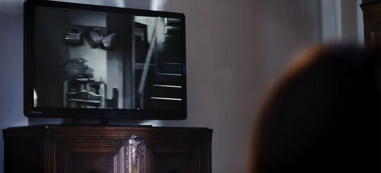The Russian Bride (2019) movie still: medium — A dark room features a flat screen television sitting atop an antique wooden cabinet. A person is…