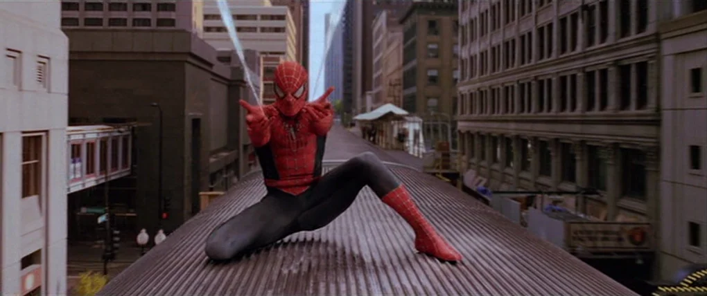 Spider-Man 2 (2004) movie still: fullbody — A character crouches on the metal roof of a moving elevated passenger train. Buildings line both sides…