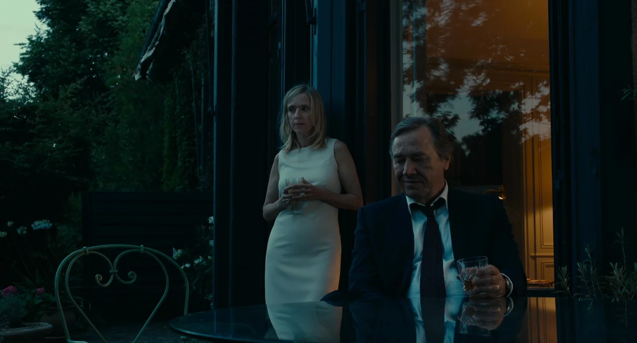 Last Summer (2023) movie still: medium — A patio area at dusk is adjacent to a softly lit indoor space. A man and a woman in formal attire stand and…
