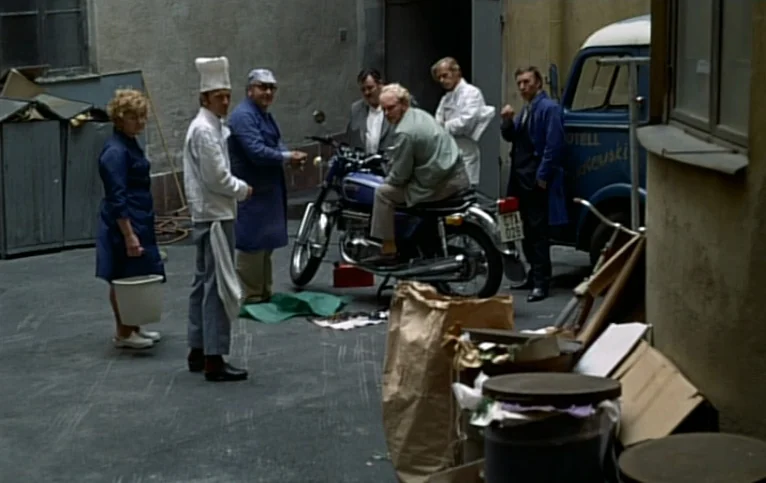 Giliap (1975) movie still: fullbody — A group of people gather in a paved courtyard near a parked motorcycle. Several men and a woman dressed in…