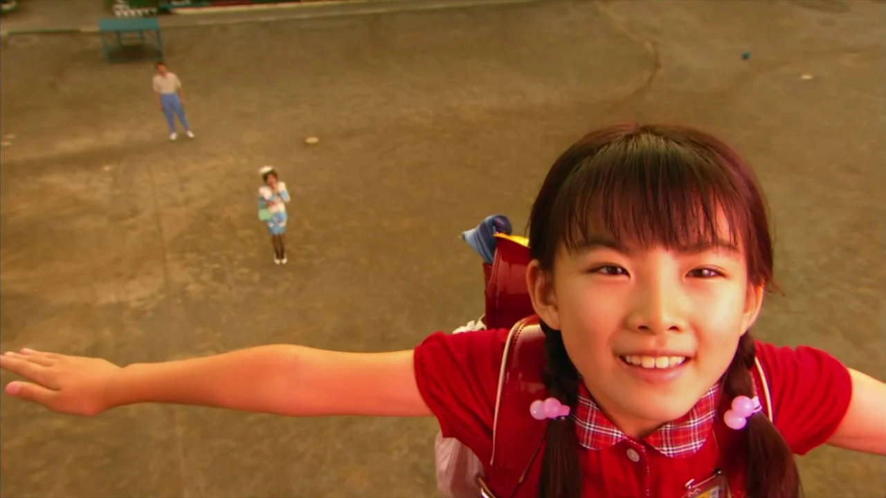 Kamikaze Girls (2004) movie still: closeup — A vast, paved schoolyard shows a young girl in the foreground with arms outstretched. Two adults stand…