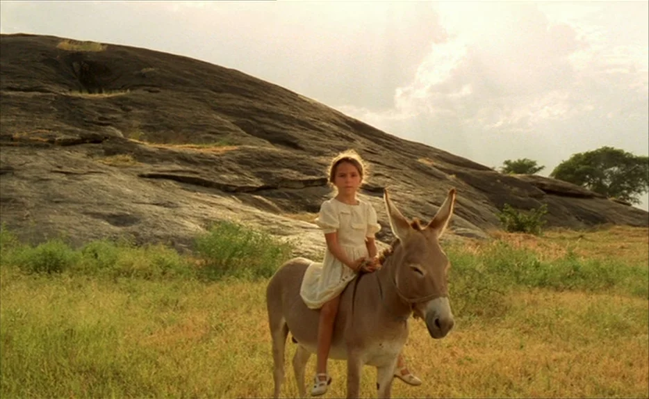 Chocolat (1988) movie still: fullbody — An open grassy field features a large, dark, smooth rock formation in the background. A young girl in a…