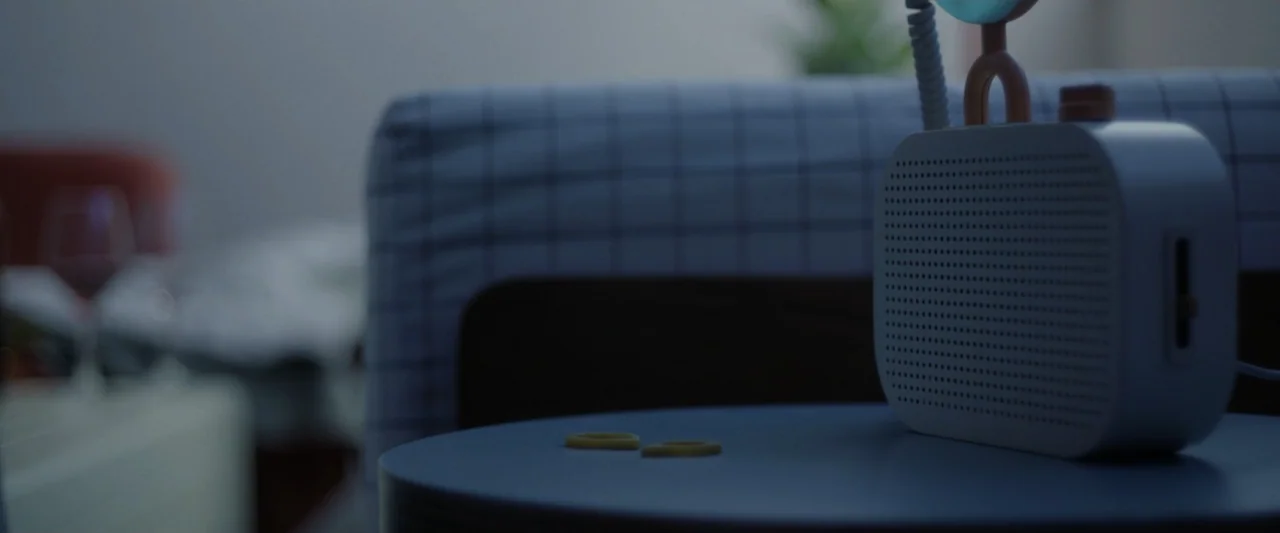 Sign Here (2017) movie still: closeup — A dark bedside table holds a white portable speaker and two small rings. The background shows a soft-focus…