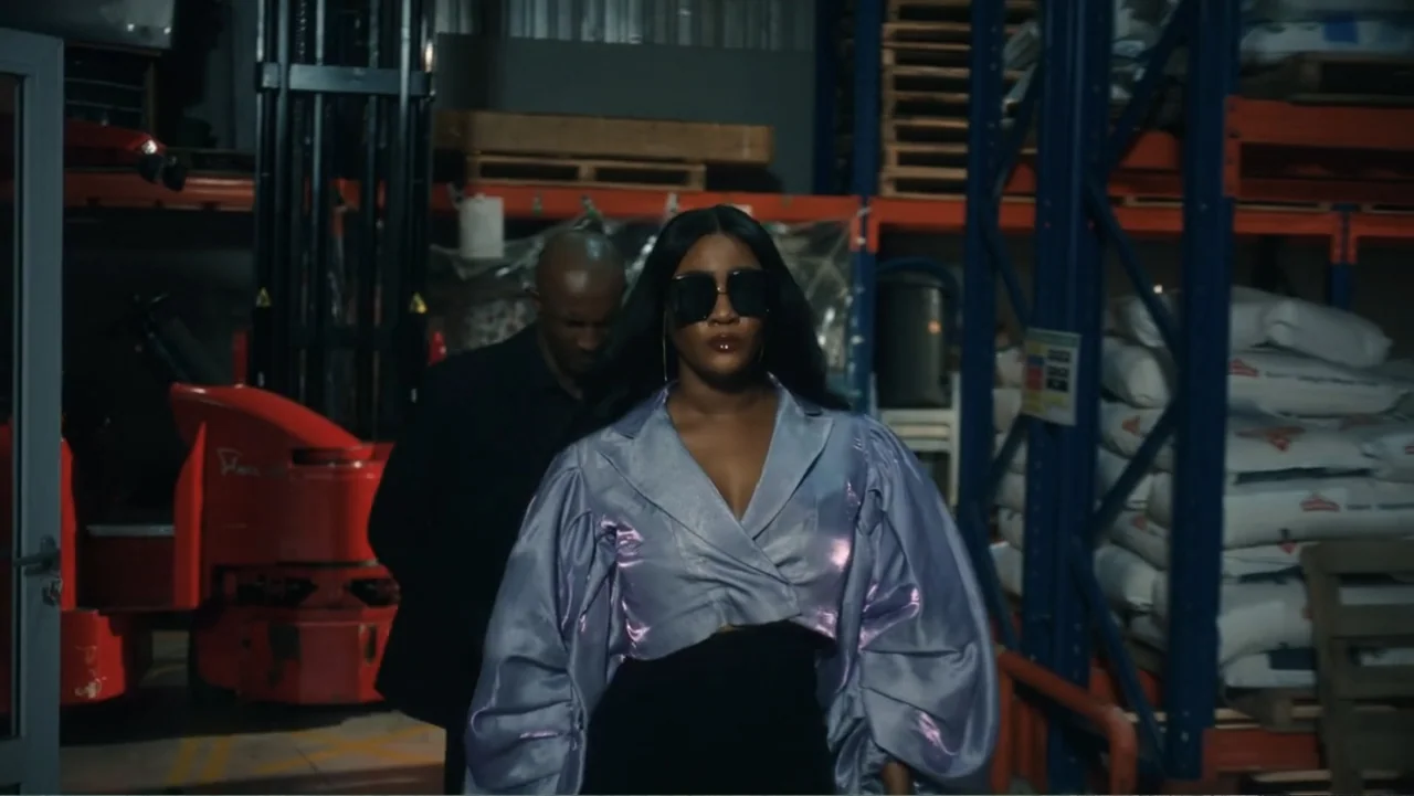 SIN: Blackout (2025) movie still: medium — A dark warehouse contains stacked palletized goods and an industrial forklift in the background. A woman…