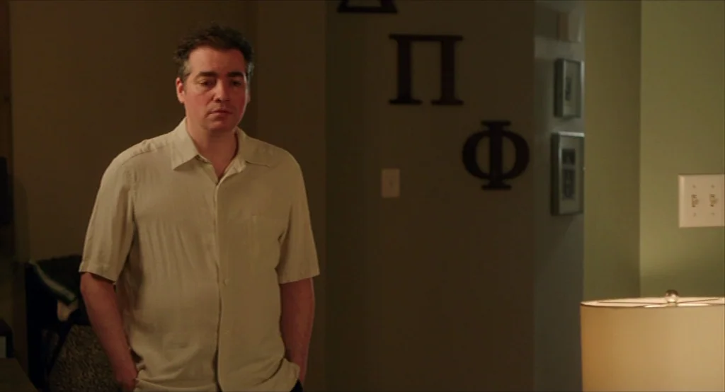 Results (2015) movie still: medium — A dimly lit room features Greek letters mounted on a sage green wall. A man in a light-colored button-up shirt…