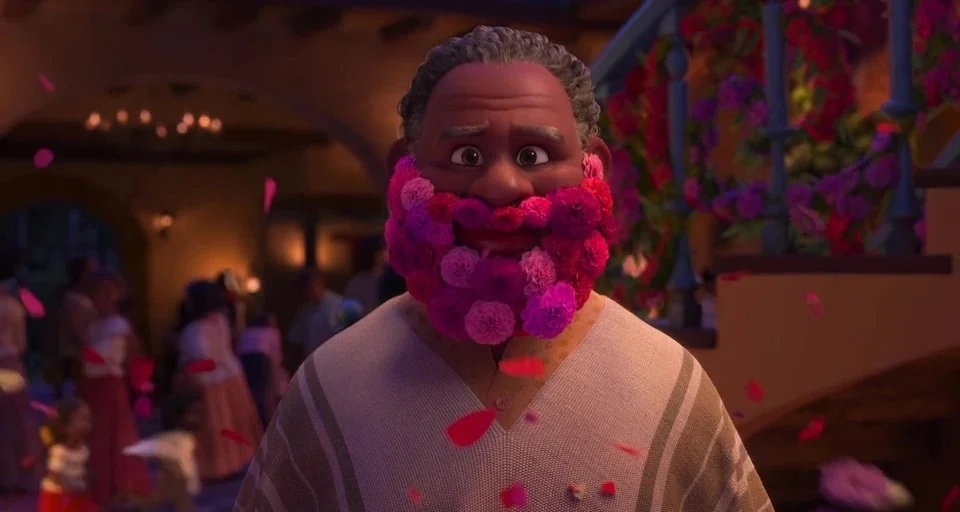 Encanto (2021) movie still: closeup — A man stands in a festive courtyard with blurred figures in the background. Pink and purple flower petals fall…