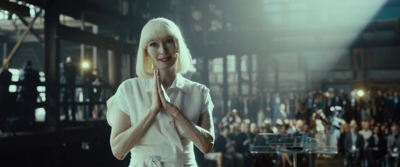 Okja (2017) movie still: medium — A spacious industrial hall with metal beams and a distant crowd of blurred people. A woman with a blonde bob…