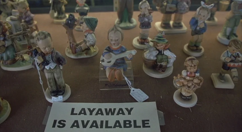 About Schmidt movie still: medium — A collection of small porcelain figurines is arranged on a flat surface. A white sign reading LAYAWAY IS…